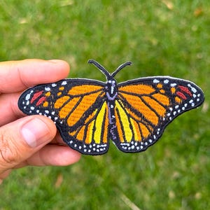 May include: Embroidered butterfly patch with a monarch butterfly design. The patch has a black border with orange, yellow, and red details. The butterfly's wings are spread, showcasing its intricate patterns.