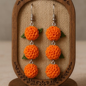 Puede incluir: Pendientes colgantes florales naranjas con detalles de hojas verdes. Los pendientes se exhiben en un soporte de madera con un dise&ntilde;o tallado decorativo. Los pendientes est&aacute;n hechos de un material texturizado y tienen ganchos plateados.