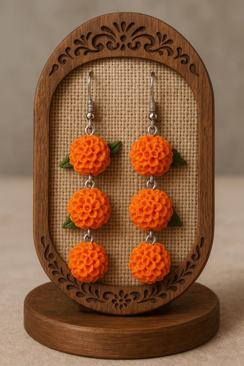 Puede incluir: Pendientes colgantes florales naranjas con detalles de hojas verdes. Los pendientes se exhiben en un soporte de madera con un dise&ntilde;o tallado decorativo. Los pendientes est&aacute;n hechos de un material texturizado y tienen ganchos plateados.