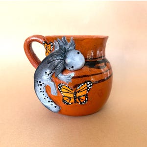 May include: A terracotta mug with a hand-painted axolotl and butterfly design. The mug has a brown glaze with black stripes and a handle.