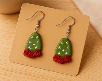 May include: A pair of handmade earrings featuring a green cactus design with white crosses and red rose accents. The earrings have silver-colored hooks and are displayed on a tan card.
