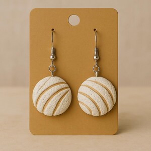 May include: A pair of white and tan earrings shaped like concha bread rolls, hanging from silver hooks. The earrings are displayed on a brown card with a hole at the top. The concha earrings are a unique accessory.