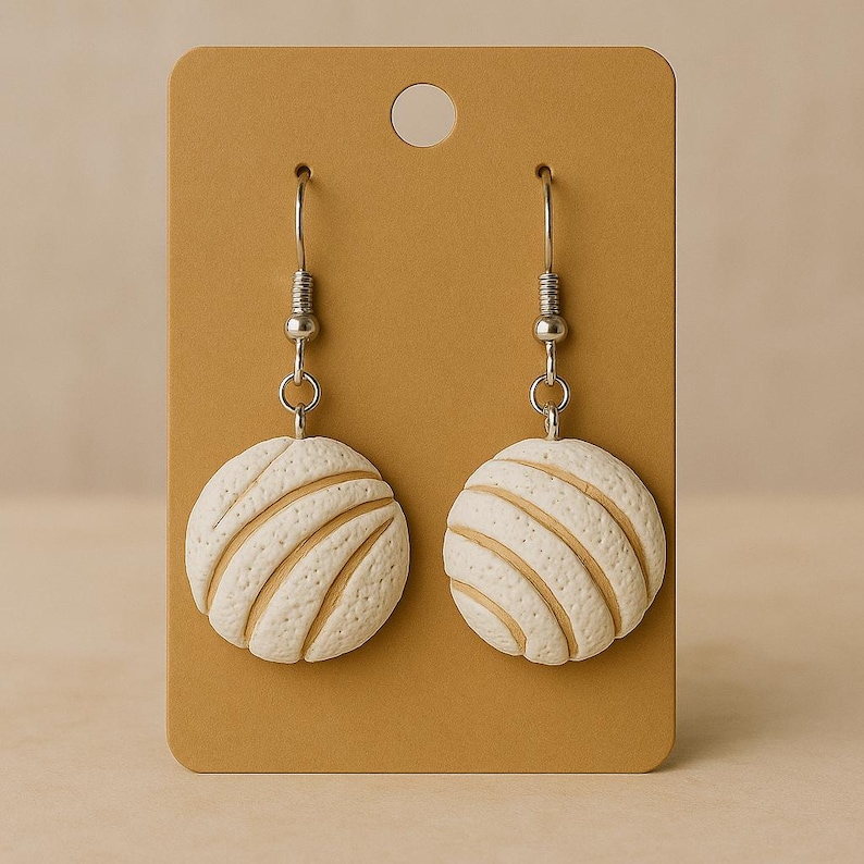 May include: A pair of white and tan earrings shaped like concha bread rolls, hanging from silver hooks. The earrings are displayed on a brown card with a hole at the top. The concha earrings are a unique accessory.