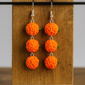Puede incluir: Un par de pendientes de bola naranja texturizados con detalles de hojas verdes. Los pendientes cuelgan de ganchos plateados y se exhiben en un soporte de madera. Los pendientes tienen un dise&ntilde;o &uacute;nico y detallado.