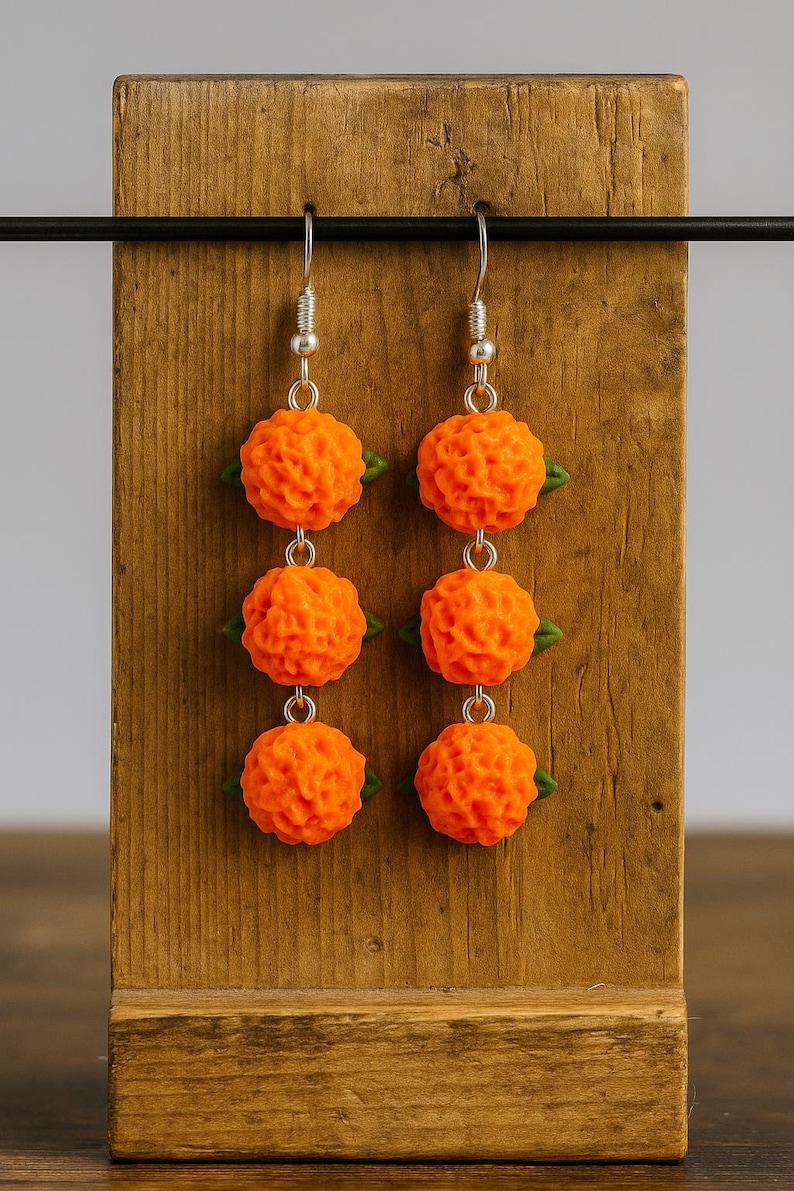 Puede incluir: Un par de pendientes de bola naranja texturizados con detalles de hojas verdes. Los pendientes cuelgan de ganchos plateados y se exhiben en un soporte de madera. Los pendientes tienen un dise&ntilde;o &uacute;nico y detallado.
