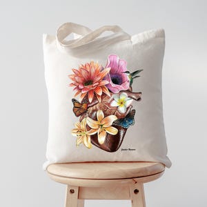 May include: A white tote bag featuring a colourful illustration of a human heart surrounded by flowers and butterflies. The flowers are pink, orange, yellow, and white. The butterflies are orange, yellow, and blue. The text "Jennifer Romero" is printed in the bottom right corner of the bag.