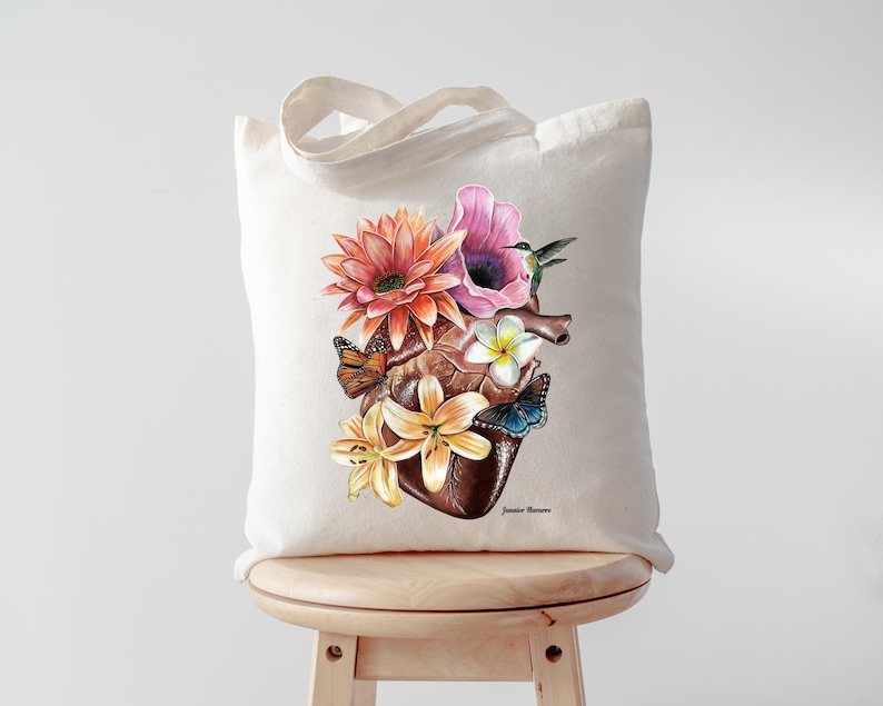 May include: A white tote bag featuring a colourful illustration of a human heart surrounded by flowers and butterflies. The flowers are pink, orange, yellow, and white. The butterflies are orange, yellow, and blue. The text "Jennifer Romero" is printed in the bottom right corner of the bag.