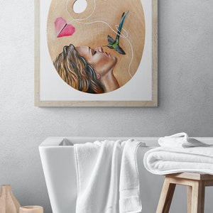 May include: A framed painting of a woman with long brown hair, looking up at a hummingbird with a pink paper heart attached to its tail by a string. The painting is on a wooden palette.