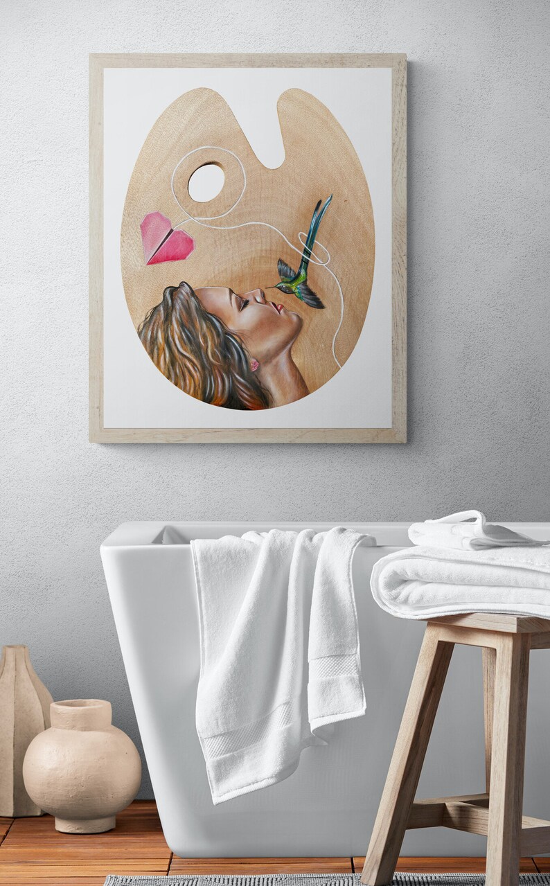 May include: A framed painting of a woman with long brown hair, looking up at a hummingbird with a pink paper heart attached to its tail by a string. The painting is on a wooden palette.