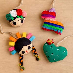 Könnte beinhalten: Vier farbenfrohe Filzornamente mit Aufhängeschnüren. Die Ornamente umfassen ein von Frida Kahlo inspiriertes Gesicht mit Blumenkranz, eine regenbogenfarbene Piñata, ein Mädchen mit Zöpfen und farbenfrohem Sonnenaufgang sowie einen herzförmigen Kaktus.