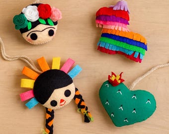 May include: Four felt hanging ornaments featuring a Frida Kahlo-inspired face with floral crown, a multi-colored fringed piñata, a character with black braided hair and a rainbow sunburst headpiece, and a green heart-shaped cactus with a red flower.