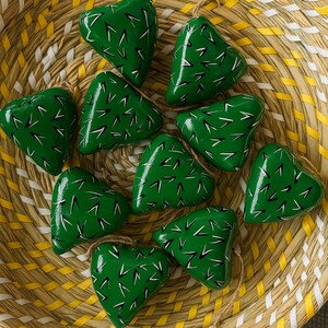 May include: A collection of ten heart-shaped ornaments with a vibrant green finish, adorned with white and black linear details, resting inside a woven basket with yellow and white stripes. Each ornament is suspended by a natural twine loop.