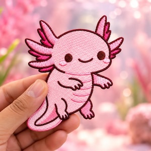 May include: An embroidered patch shaped like a light pink axolotl with dark pink gills, a smiling face, and small limbs. The patch features a dark pink border outlining the entire figure, with detailed stitching throughout the body and facial features.