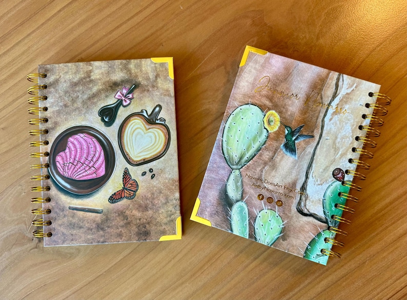 May include: Two spiral bound notebooks with colorful illustrations. The first notebook features a pink heart-shaped pastry, a butterfly, and a coffee cup with a heart-shaped design. The second notebook features a hummingbird, a cactus, and the text "Navajo Dreams Art Fry Co. Art by Marjoria Kim".