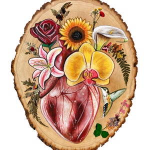 May include: A painted wooden slice with a realistic anatomical heart surrounded by flowers, including a sunflower, a lily, an orchid, and a calla lily. A hummingbird is perched on the heart, with its beak touching the orchid. The background is a light brown wood.