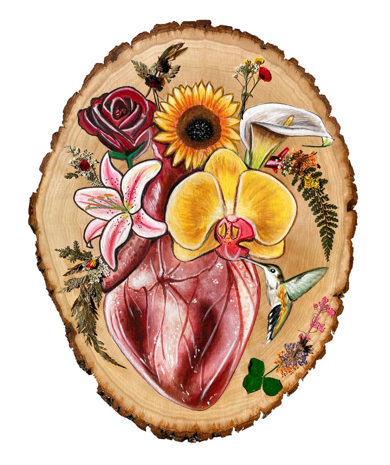 May include: A painted wooden slice with a realistic anatomical heart surrounded by flowers, including a sunflower, a lily, an orchid, and a calla lily. A hummingbird is perched on the heart, with its beak touching the orchid. The background is a light brown wood.