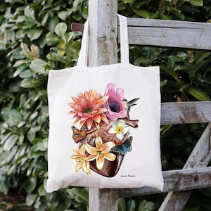 May include: A white tote bag featuring a colourful illustration of a human heart surrounded by flowers and butterflies. The heart is brown and the flowers are pink, yellow, orange, and white. The butterflies are orange, blue, and brown. The text "Jamie Rae Murray" is printed in black at the bottom of the bag.