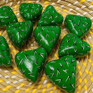 May include: A collection of heart-shaped, green painted stones with white and black markings, arranged in a woven basket. The basket has a natural, light brown color with yellow and white accents. The stones appear to be decorative.