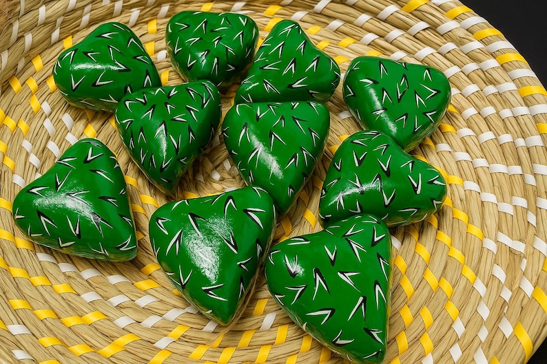 May include: A collection of heart-shaped, green painted stones with white and black markings, arranged in a woven basket. The basket has a natural, light brown color with yellow and white accents. The stones appear to be decorative.