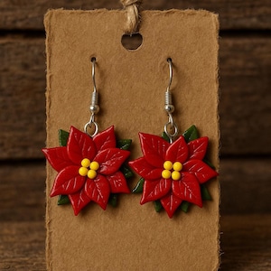 May include: A pair of poinsettia earrings. Each earring features a vibrant red flower with yellow centers and green leaves. The earrings are displayed on a brown card with a jute string for hanging. The background is a wooden surface.