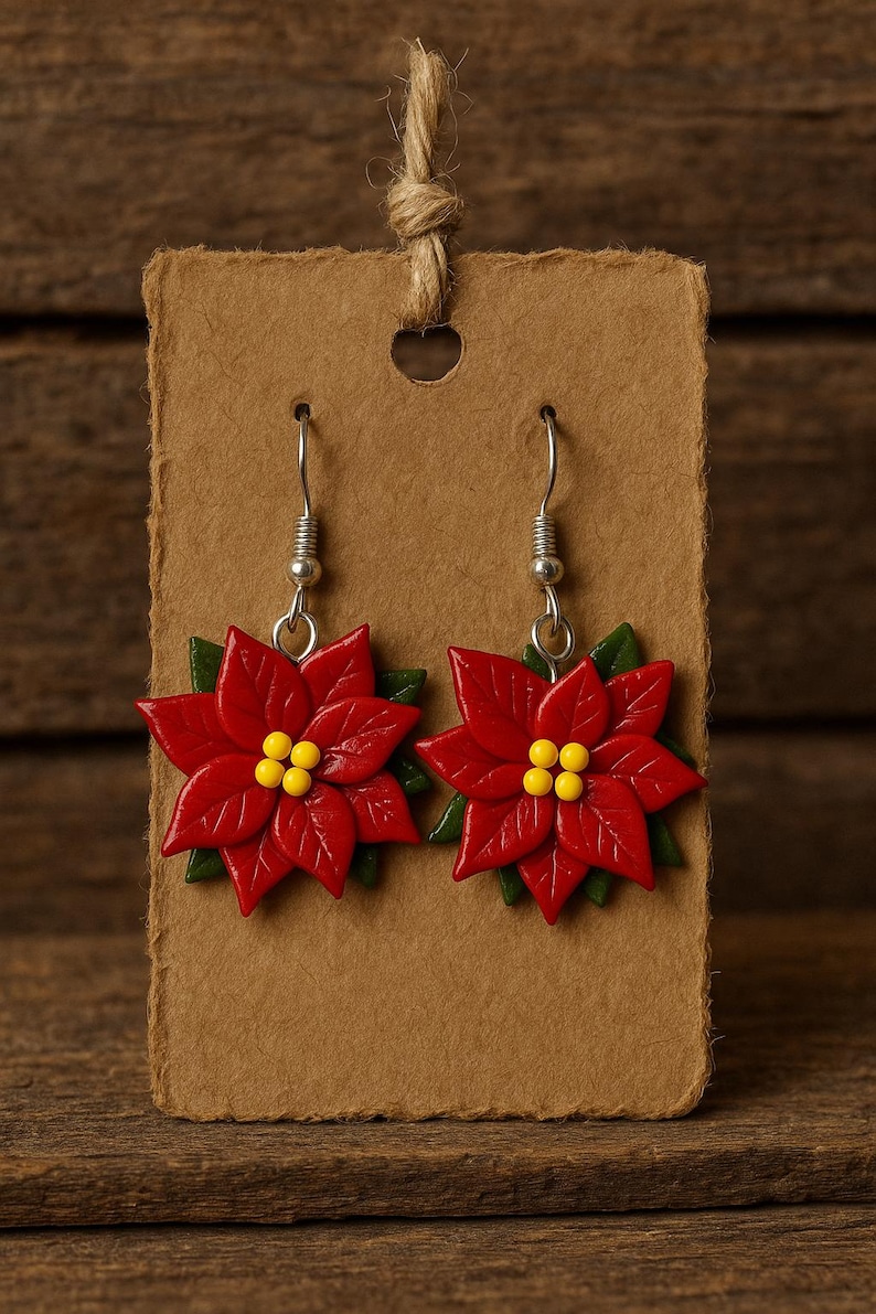 May include: A pair of poinsettia earrings. Each earring features a vibrant red flower with yellow centers and green leaves. The earrings are displayed on a brown card with a jute string for hanging. The background is a wooden surface.