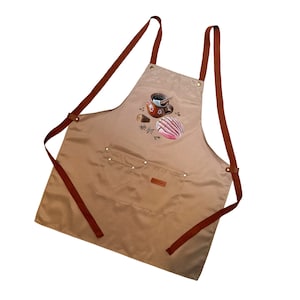 May include: Tan apron with brown straps and a front pocket. The apron features a colorful illustration of a cup of coffee, a concha pastry, and other coffee-related items. The apron is designed for kitchen use.