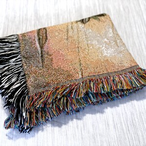 May include: A woven throw blanket with a colourful fringe border. The blanket features a tapestry-like design with shades of brown, beige, and orange.