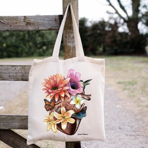 May include: A white tote bag featuring a colourful floral design with a heart, butterflies, and a hummingbird. The design is centred on the bag and includes the text "Jamie Moore".
