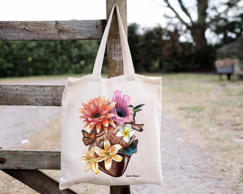 May include: A white tote bag featuring a colourful floral design with a heart, butterflies, and a hummingbird. The design is centred on the bag and includes the text "Jamie Moore".