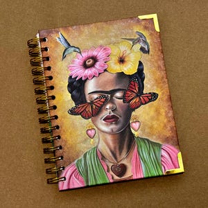 May include: A spiral bound journal with a colorful cover featuring a painting of Frida Kahlo with butterflies over her eyes and flowers in her hair. The background is a golden yellow.