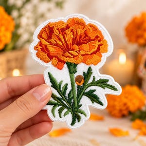 Puede incluir: Un parche bordado que presenta una flor de cempasúchil de color naranja y amarillo vibrante con un tallo y hojas verdes. El diseño se sitúa sobre un fondo blanco con un borde troquelado que sigue la forma de la ilustración floral.
