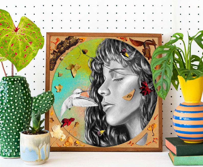 May include: A framed mixed media artwork featuring a black and white portrait of a woman with her eyes closed. The woman's face is surrounded by a circle of dried flowers and leaves, including a hummingbird in flight. The artwork has a rustic, natural feel.