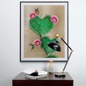 May include: A framed print of a heart-shaped cactus with pink flowers and a butterfly. The print is hanging on a white wall above a wooden table with a lamp and a vase.