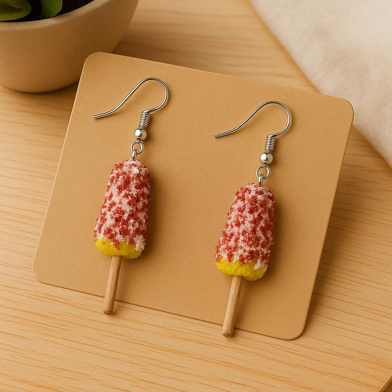 May include: Novelty earrings with popsicle charms. Each earring has a silver hook, a popsicle-shaped charm with a red and white speckled top, a yellow base, and a wooden stick. The earrings are displayed on a tan card.