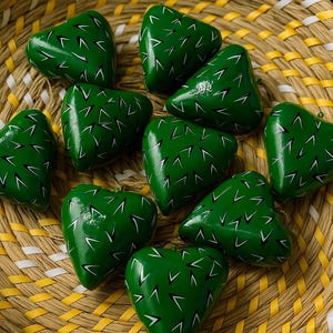 May include: A collection of green heart-shaped ornaments with white accents, resembling cacti, are arranged in a woven basket. The basket has a yellow and white pattern. The ornaments are likely handmade and decorative.