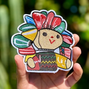 May include: Embroidered patch featuring a colorful, smiling, traditional Mexican doll with a red, green, and yellow headdress. The doll is wearing a black and green patterned shirt and holding a yellow object.