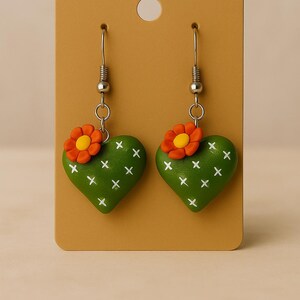 May include: A pair of green heart-shaped earrings with white cross details, resembling cacti. Each earring features a small orange flower with a yellow center. The earrings hang from silver-colored hooks and are displayed on a tan card.