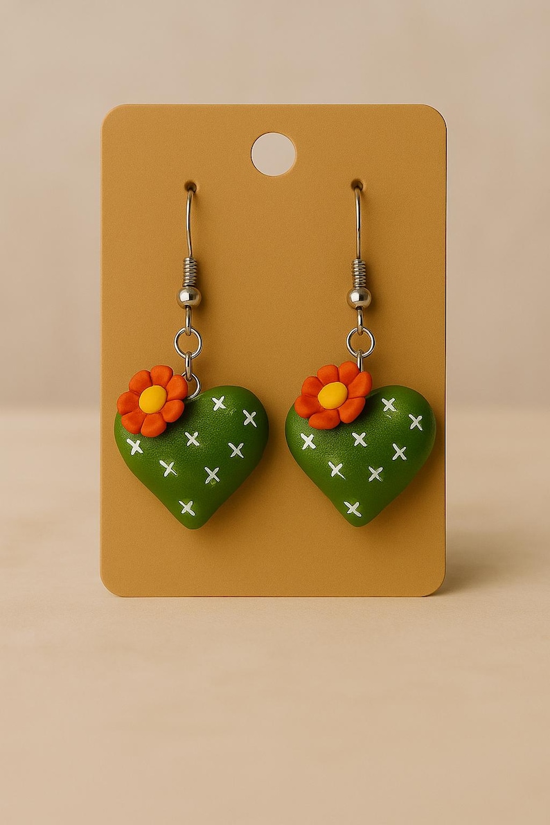 May include: A pair of green heart-shaped earrings with white cross details, resembling cacti. Each earring features a small orange flower with a yellow center. The earrings hang from silver-colored hooks and are displayed on a tan card.