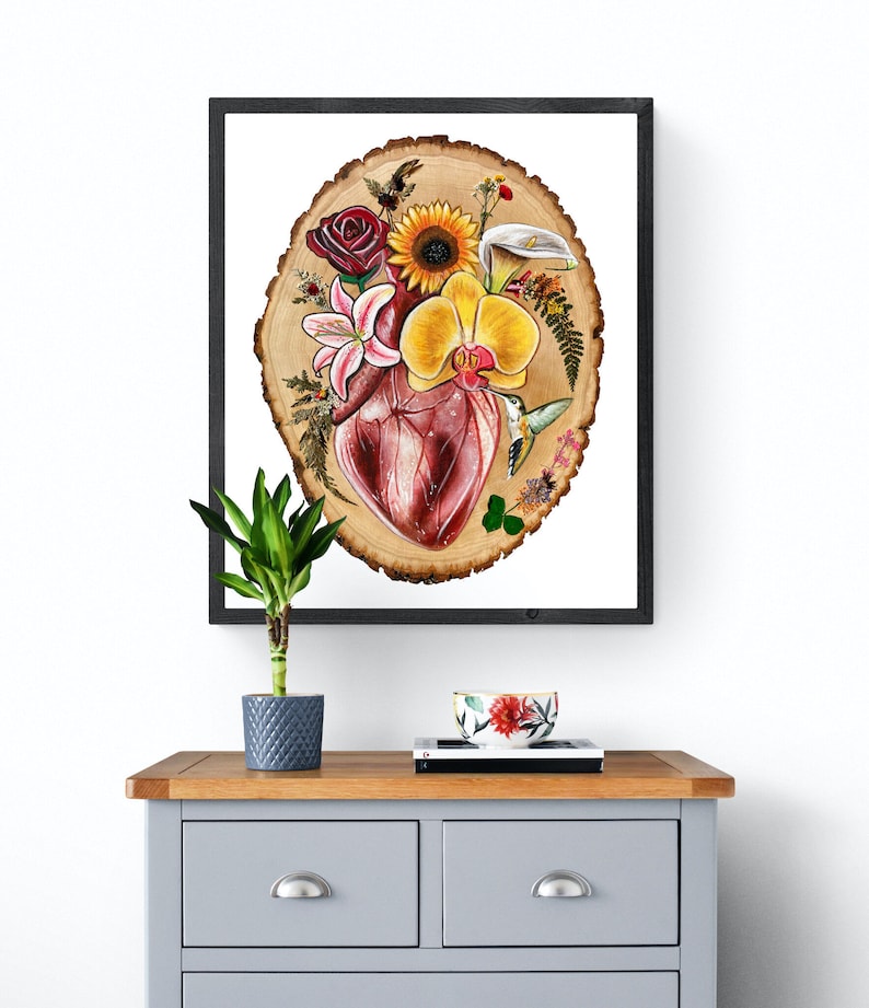 May include: A framed print of a watercolor painting depicting a human heart surrounded by flowers and a hummingbird on a wood slice background.