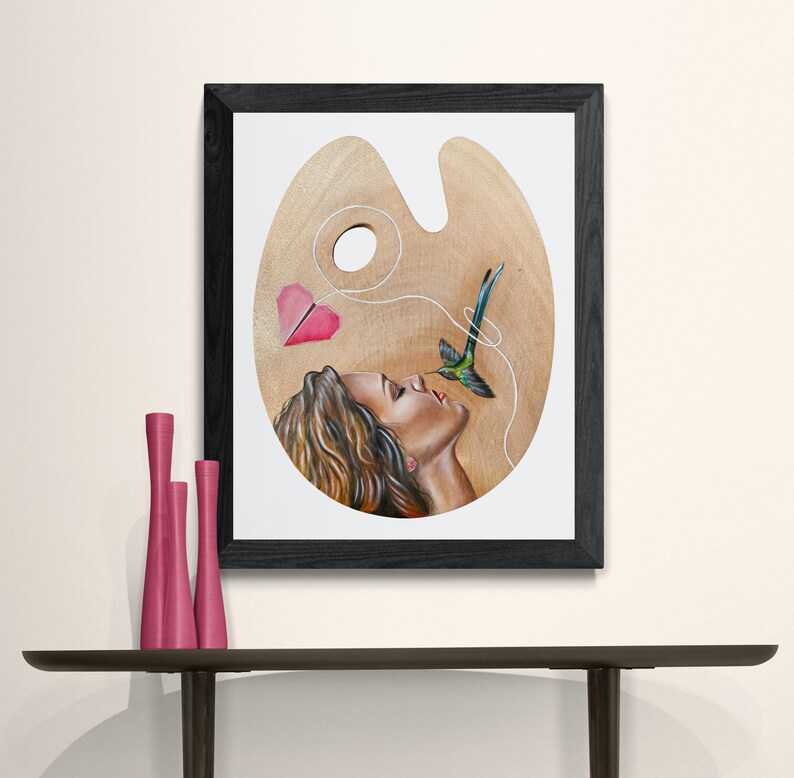 May include: A painting of a woman's face on a wooden artist's palette. The woman is looking up at a hummingbird with a pink heart-shaped paper airplane tied to its beak. The hummingbird is flying towards the woman's face.