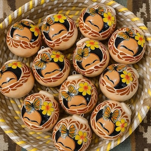 Puede incluir: Colección de adornos de cerámica pintados a mano con diseños de mariposas y flores, expuestos en una cesta tejida. Los adornos presentan detalles en naranja, negro y amarillo. La cesta tiene un asa trenzada.