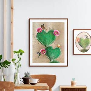 May include: Two framed illustrations of a heart-shaped cactus with pink flowers and a hummingbird. The larger frame is brown and the smaller frame is wood-toned.