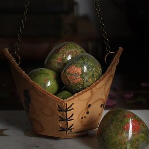 May include: A leather pouch with a chain hanging from it, filled with green and red jasper stones. The pouch is sitting on a white marble surface.