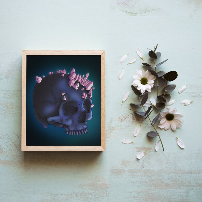 May include: A framed illustration of a dark blue skull with pink crystals on top. The skull is set against a dark blue background. The frame is made of wood and has a light brown finish.