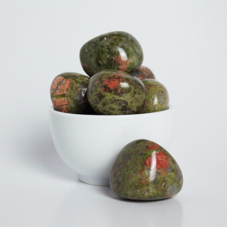 May include: A white bowl filled with green and red tumbled stones. The stones are smooth and have a speckled appearance.