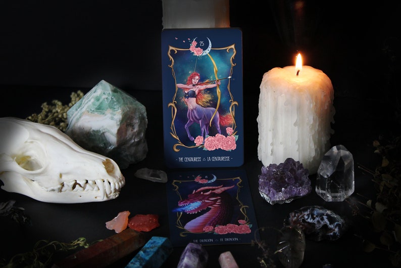 May include: A tarot card reading set with a candle, crystals, and a skull. The card in the foreground shows a centaur woman with a bow and arrow, with the text "The Chariot - La Chariote" on the card.