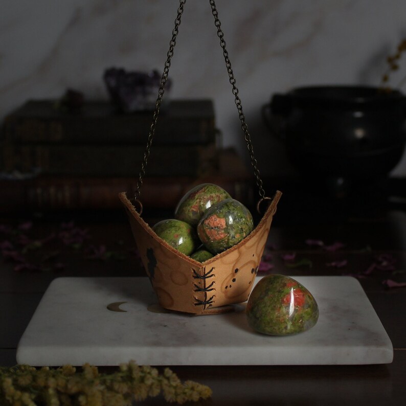 May include: A leather pouch with a chain hanging from it, filled with green and red jasper stones. The pouch is sitting on a white marble surface.