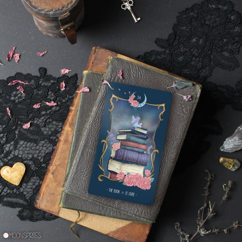 May include: A blue tarot card with a crescent moon and a fairy sitting on a stack of books. The card is titled "The Book & Le Livre" and has the number 34 on it. The card is surrounded by pink flowers and a dark blue background.