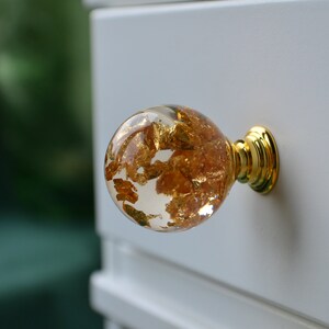 May include: A round, clear glass cabinet knob with gold leaf accents and a gold metal base.