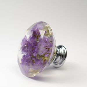 May include: A decorative knob with a silver base, featuring a purple flower encased in clear resin.